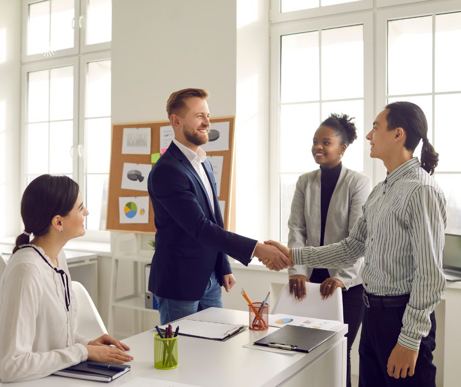 4 people around a table, one man is standing and shaking hands with a couple that is facing him.