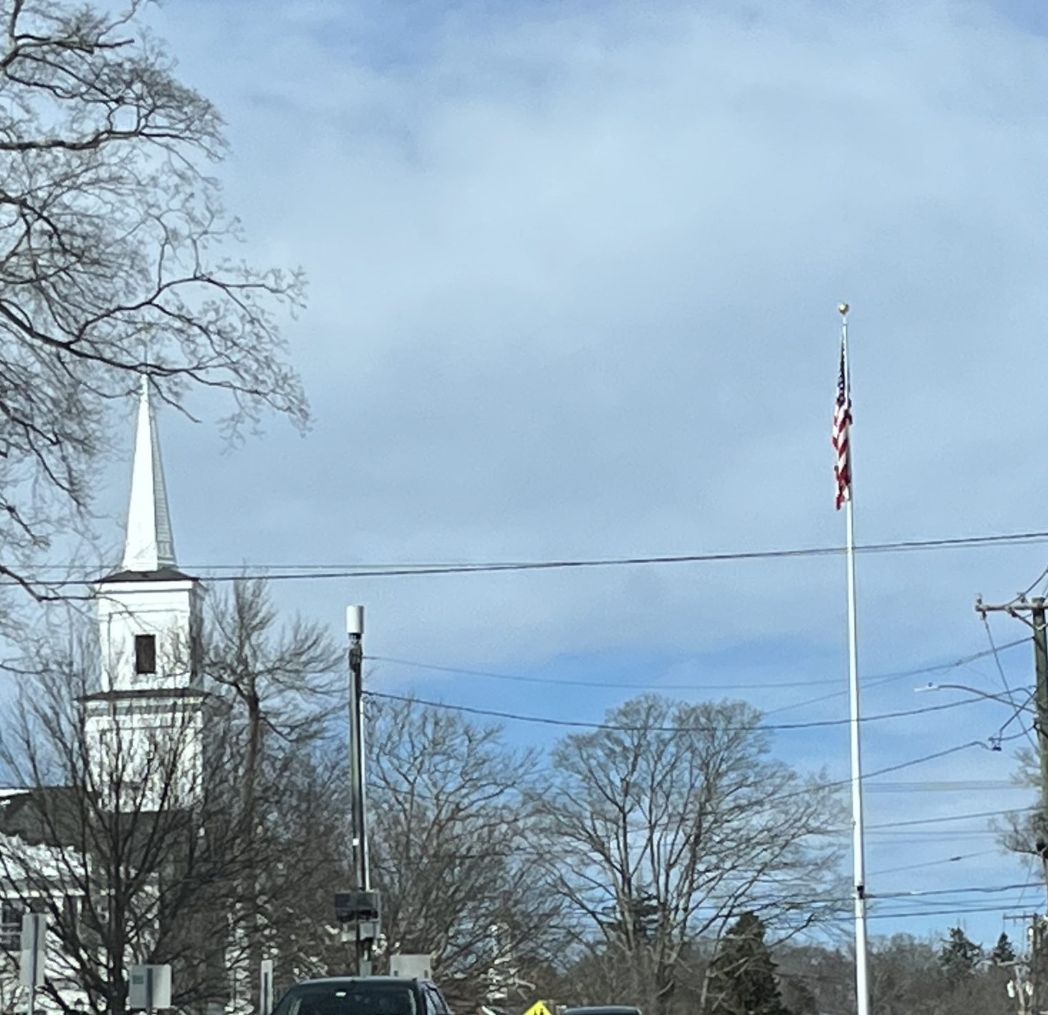Picture of open sky with a white clabboard church spire on the left and an American flag on the right