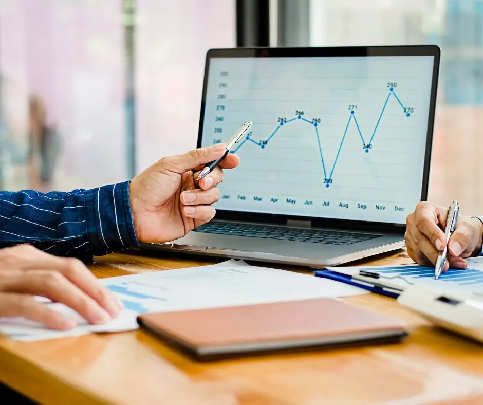 Picture is of a computer screen showing an upward trending graph and a hand pointing to the computer